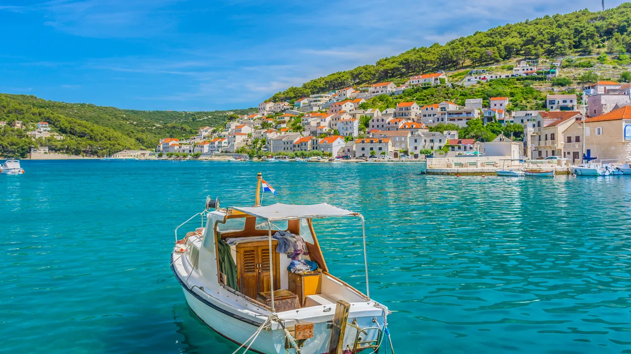 A small boat at anchor in a turquoise bay below a hillside village on the island of Brač