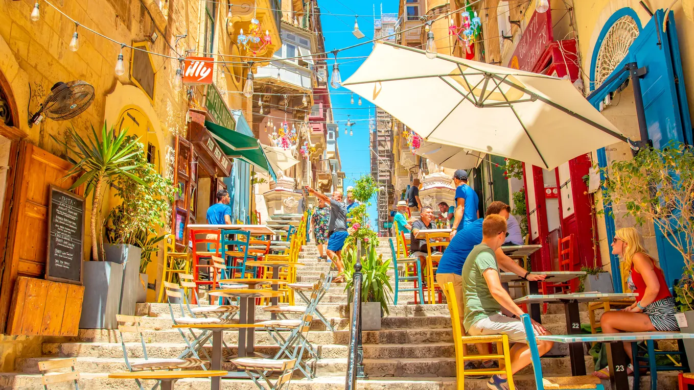 A sunlit stepped street of warm limestone buildings, balconies and café tables in Valletta’s old town