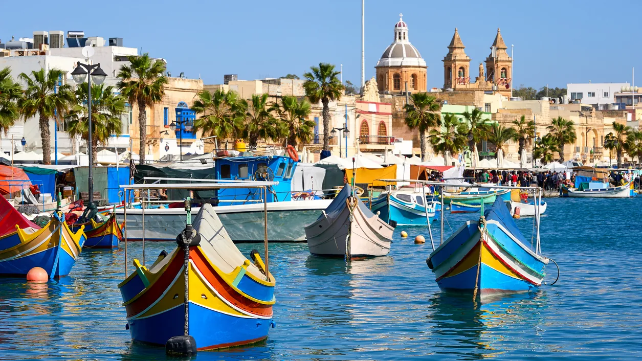 Brightly painted traditional luzzu fishing boats in Marsaxlokk harbour, with the parish church on the waterfront